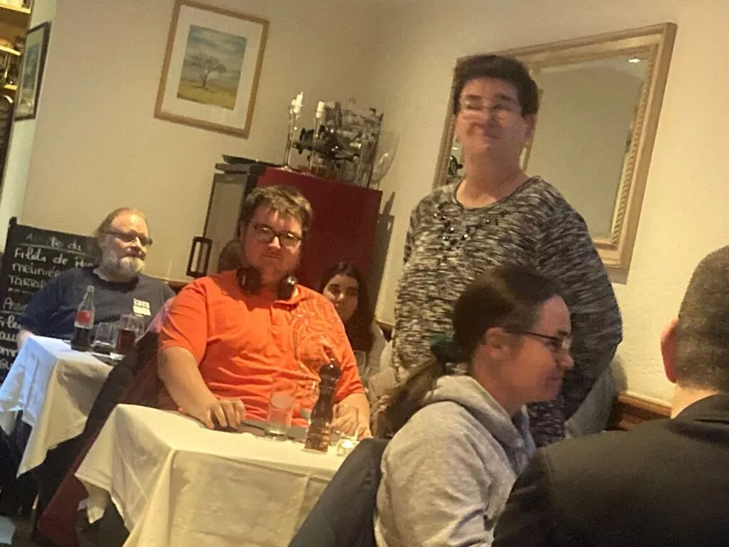 Groupe de personnes rassemblées dans un restaurant avec une femme debout au premier plan et des invités assis en conversation.