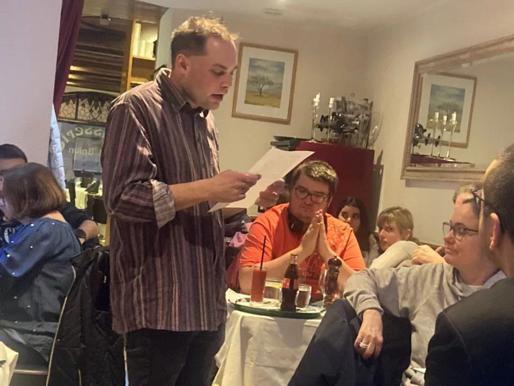Homme lisant à haute voix un journal dans un restaurant avec des spectateurs.