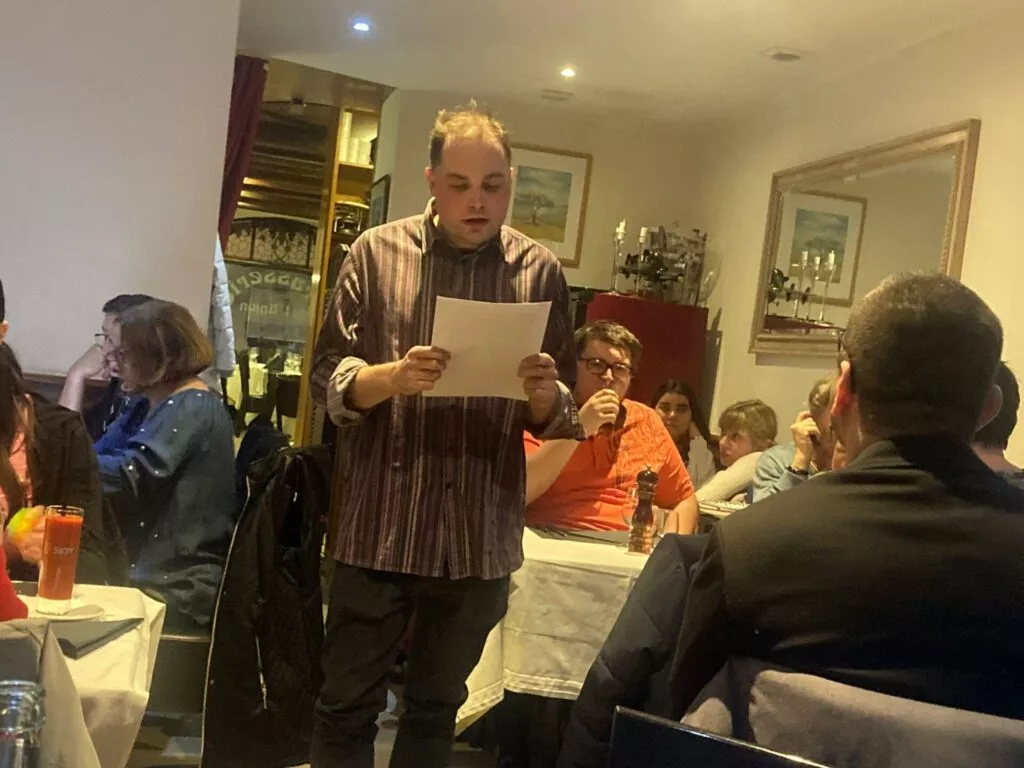 Homme debout et lisant un journal dans un restaurant avec des invités assis.