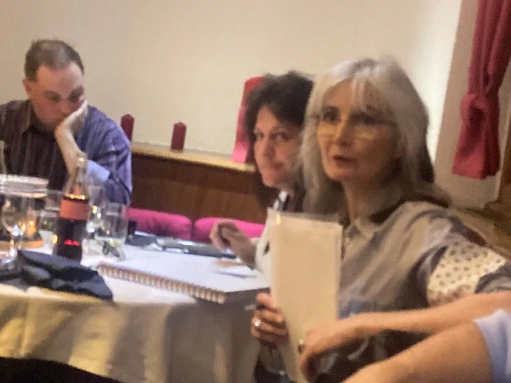 Trois individus lors d'un rassemblement intérieur, deux femmes et un homme, assis à une table avec des papiers et des boissons, semblant engagés dans une discussion ou une présentation.