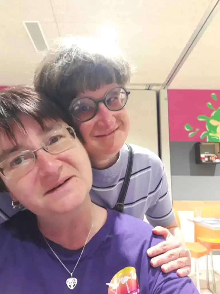 Deux femmes posent ensemble à l'intérieur, l'une portant des lunettes et une chemise à rayures violettes et blanches, et l'autre portant également des lunettes et une chemise violette au motif coloré. Ils sourient à la caméra.