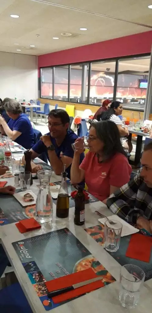 Un groupe de personnes assises à une table de restaurant avec diverses boissons et produits alimentaires, conversant et dégustant un repas. Le cadre comprend de grandes fenêtres et un intérieur coloré.