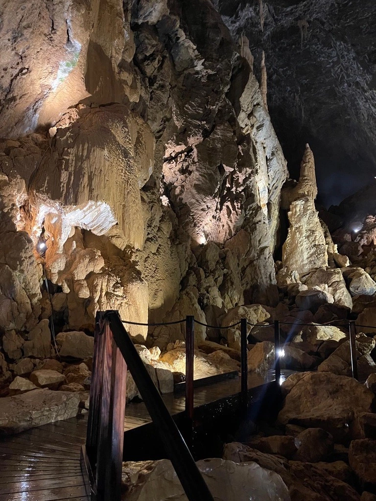 Un intérieur de grotte faiblement éclairé avec des formations rocheuses accidentées et une passerelle en bois éclairée par des lumières.