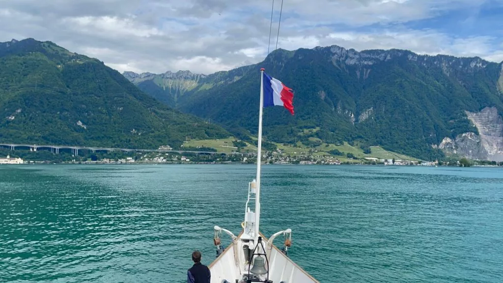 Bateau avec un drapeau français sur un lac, entouré de montagnes verdoyantes et d'un ciel partiellement nuageux.