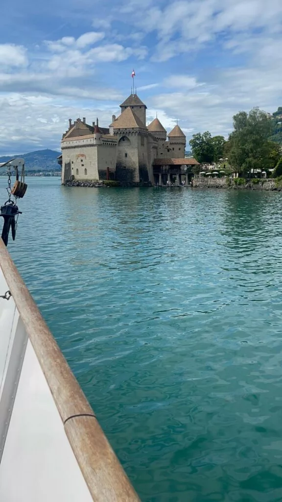 Un château au bord d'un lac calme, vu depuis un bateau, sous un ciel partiellement nuageux.