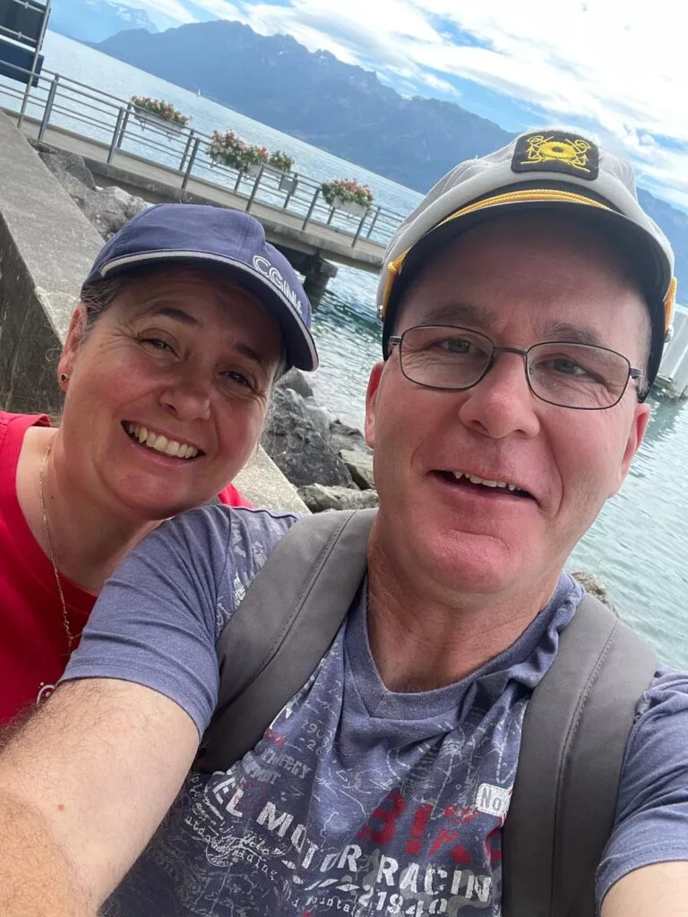 Un homme et une femme sourient pour un selfie au bord de l'eau avec des montagnes et un ciel nuageux en arrière-plan.