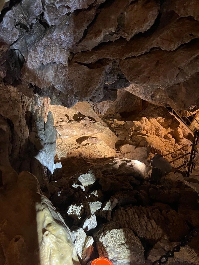Vue de l'intérieur d'une grotte avec des formations rocheuses et des stalactites, éclairée par une lumière artificielle. Une balustrade métallique est visible sur le côté droit.