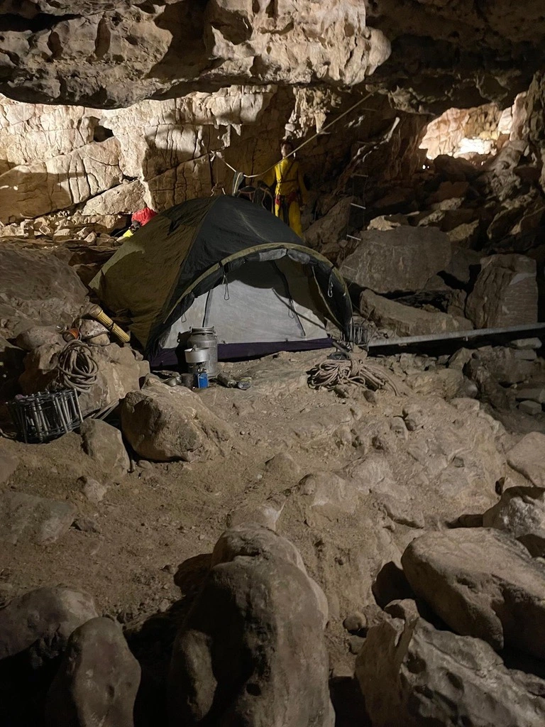 Tente installée à l'intérieur d'une grotte rocheuse, avec matériel d'escalade et lumière éclairant la zone.