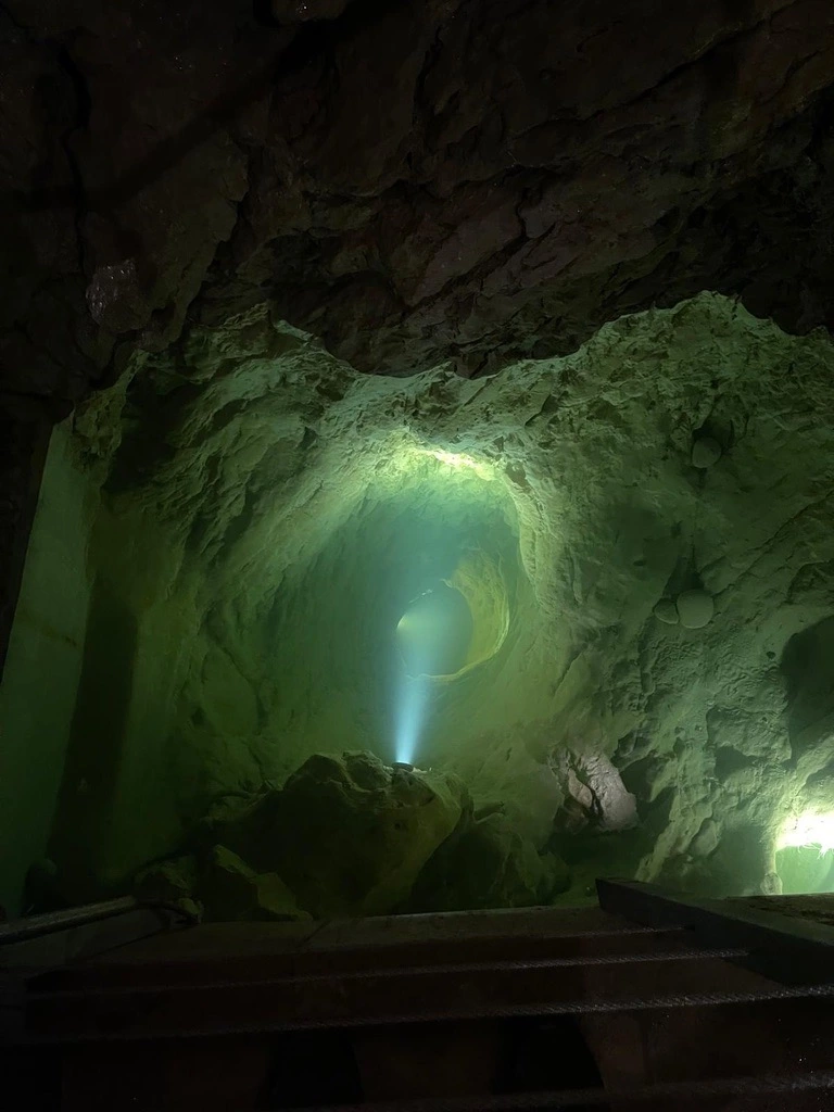 Un rayon de lumière illumine une caverne aux parois rocheuses rugueuses et à l'ouverture circulaire située au-dessus. La surface de la pierre est texturée et illuminée d'une teinte verte.