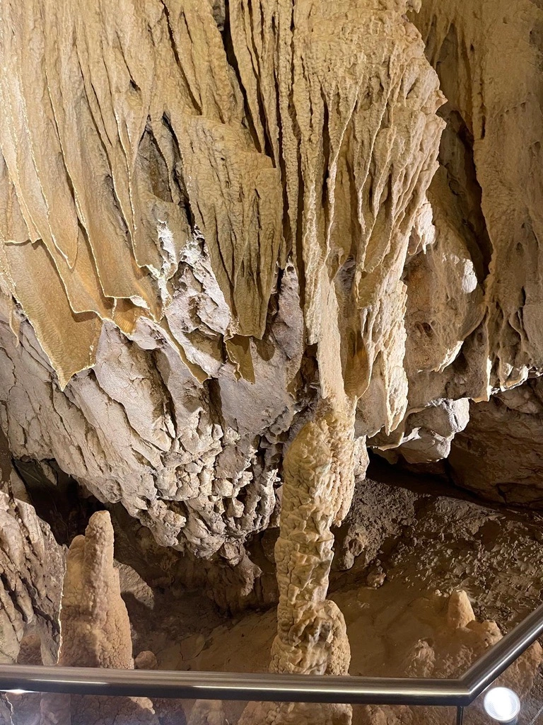 L'intérieur d'une grotte présente des stalactites suspendues au plafond et des stalagmites s'élevant du sol, avec une balustrade métallique au premier plan.