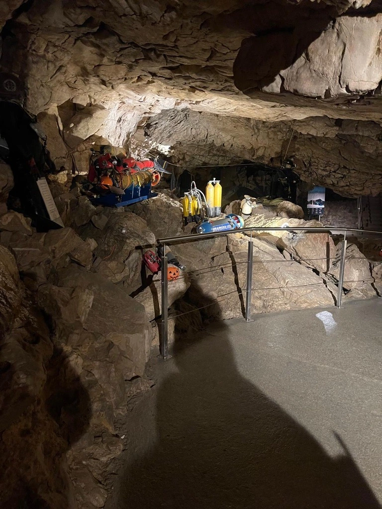 Une grotte rocheuse avec du matériel de plongée sous-marine, notamment des bouteilles et des casques, disposés sur des étagères le long du mur. Une balustrade en métal borde le passage en pierre à l'intérieur de la grotte.