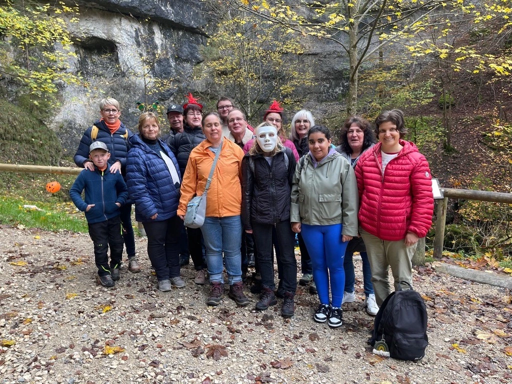 Un groupe de personnes se tient dehors sur un chemin de gravier, avec un arrière-plan rocheux et boisé. Certains portent des vestes décontractées et une personne est en costume. Il y a des feuilles d'automne sur le sol.