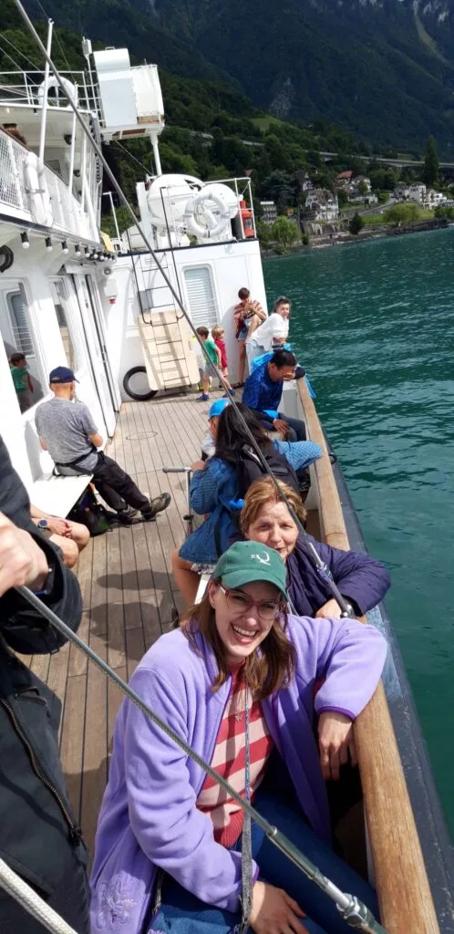 Des gens assis et debout sur le pont d'un bateau, souriant et profitant de la vue. L'arrière-plan montre un plan d'eau et un paysage montagneux.