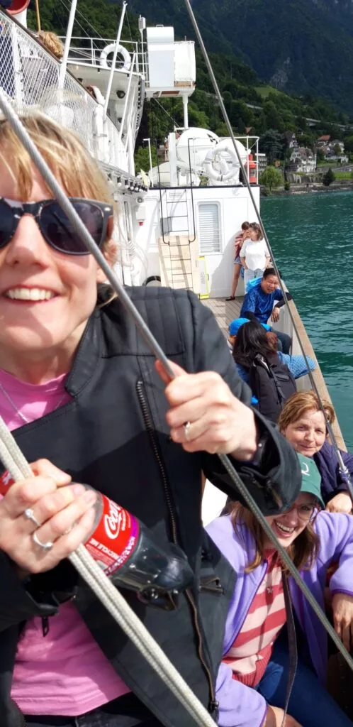 Une personne portant des lunettes de soleil sourit tout en tenant une corde sur un bateau. D'autres sont assis sur le côté. Une bouteille de Coca-Cola est visible. L'arrière-plan montre un plan d'eau et de la verdure.