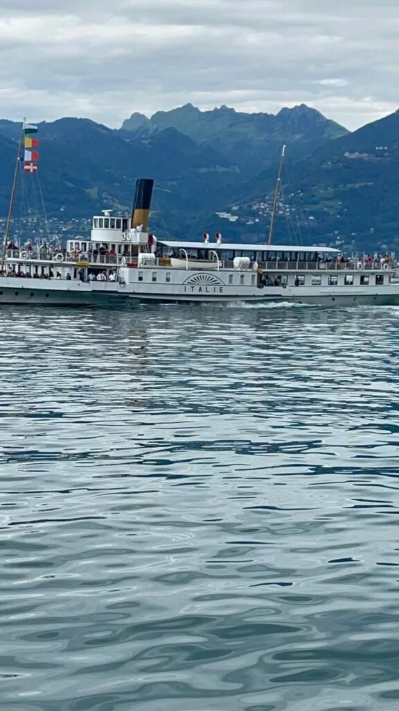 Un bateau à vapeur historique avec des drapeaux italiens navigue sur un lac calme, avec des montagnes en arrière-plan et un ciel nuageux au-dessus.