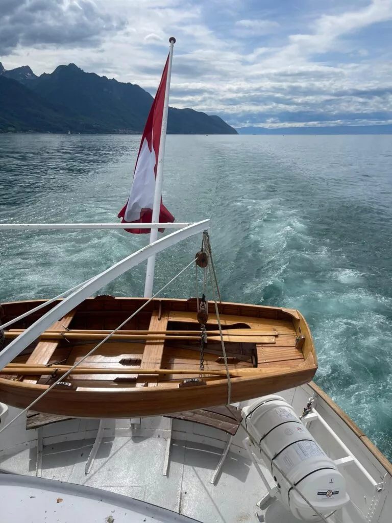 Bateau avec un drapeau suisse et un canot en bois sur le pont, naviguant sur un lac avec des montagnes et un ciel nuageux en arrière-plan.
