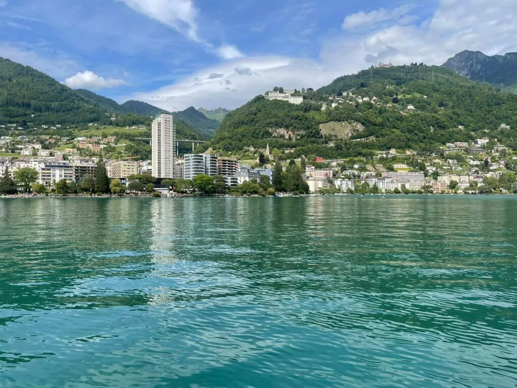 Vue sur le bord du lac d'une ville avec des collines verdoyantes en arrière-plan, avec de nombreux bâtiments, dont un grand gratte-ciel. L'eau au premier plan est calme, sous un ciel partiellement nuageux.