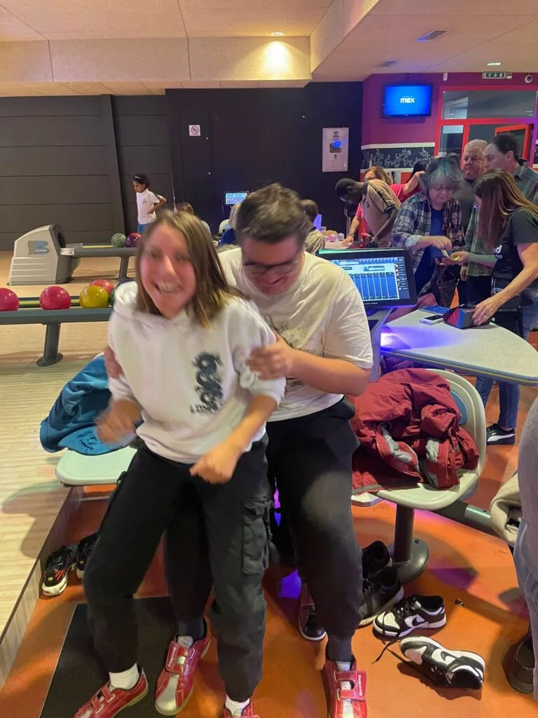 Deux personnes assises de manière ludique sur un banc dans un bowling, entourées de chaussures de bowling et d'amis en arrière-plan.