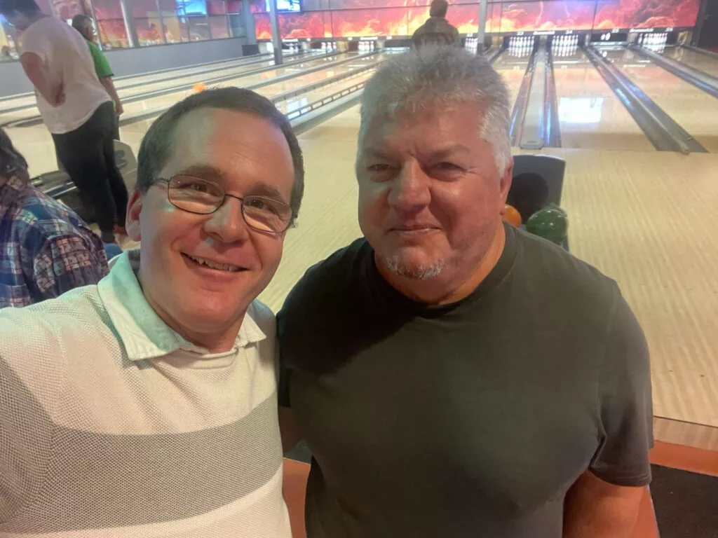 Deux hommes debout ensemble devant une piste de bowling, souriant à la caméra.