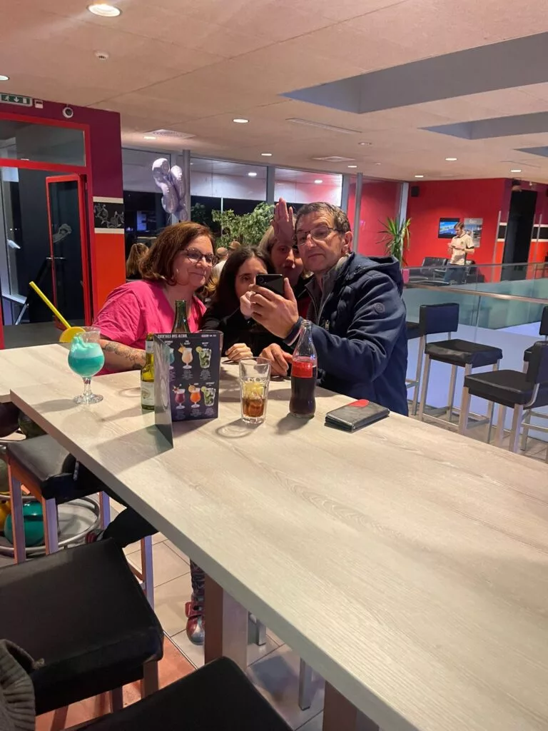 Un groupe de quatre personnes prenant un selfie à une table dans un café, avec des boissons sur la table et une décoration murale colorée en arrière-plan.