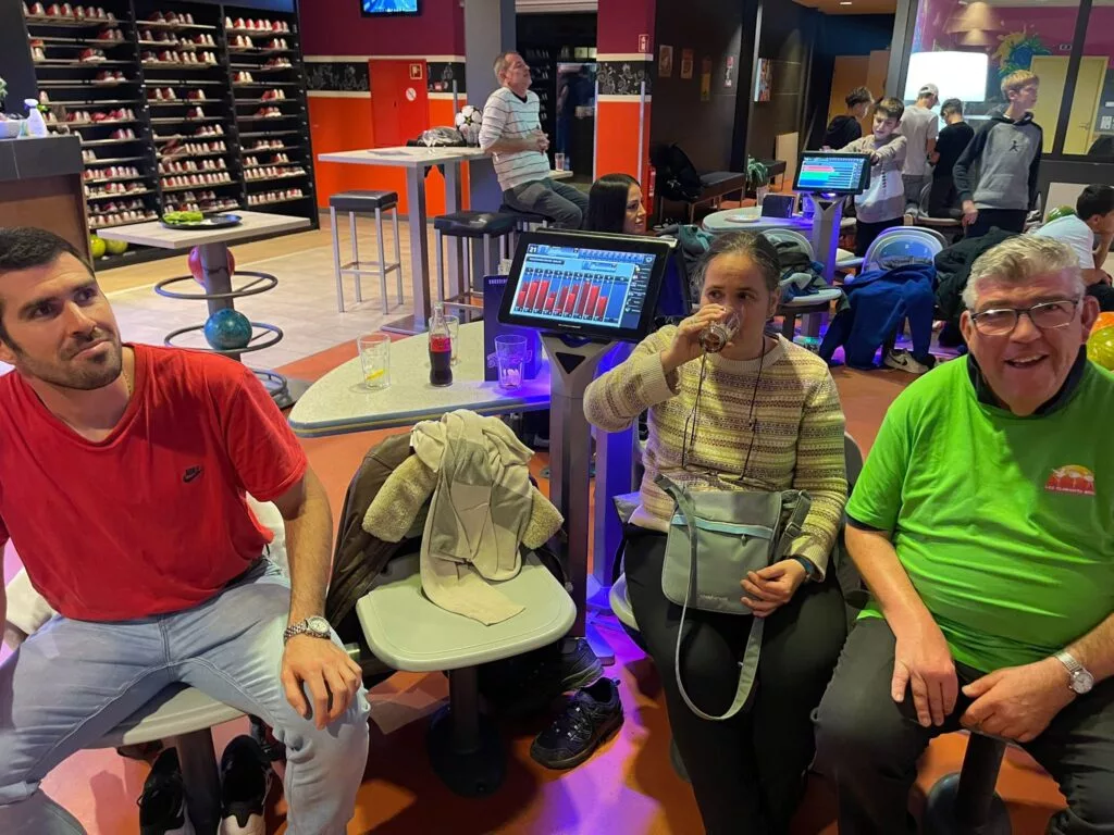 Trois personnes assises dans une salle de bowling, avec les scores visibles sur un écran derrière elles. Le décor est animé avec d'autres clients en arrière-plan.