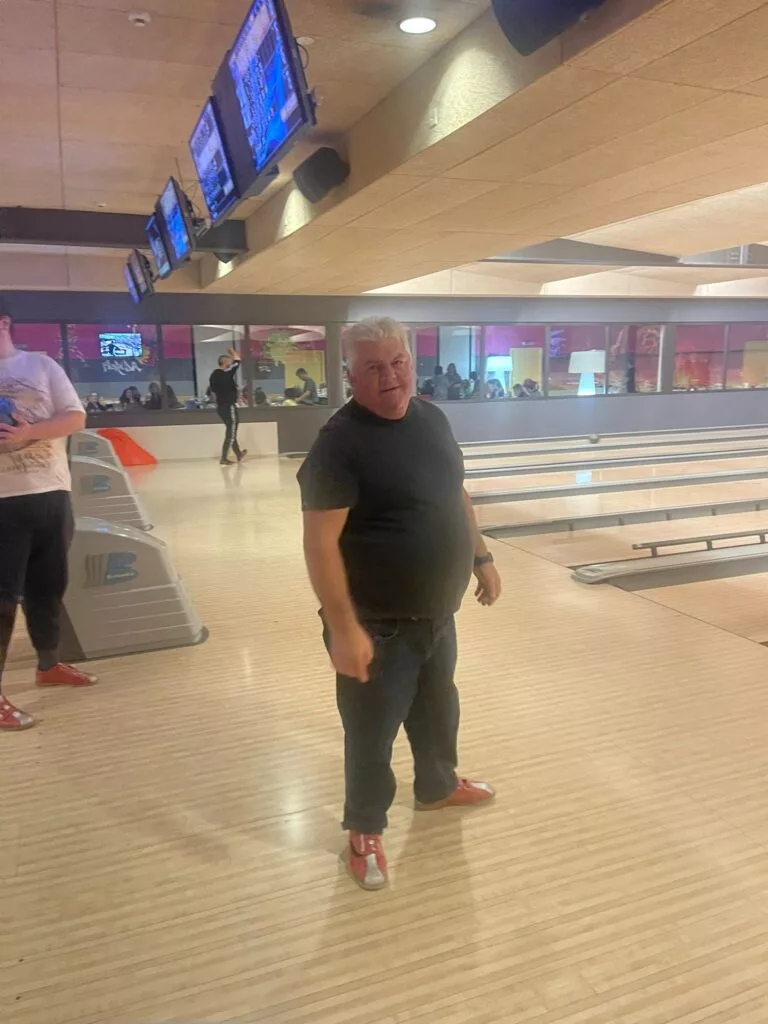 Un homme portant un t-shirt noir et un jean se tient sur une piste de bowling, avec des boules de bowling et des pistes visibles en arrière-plan.