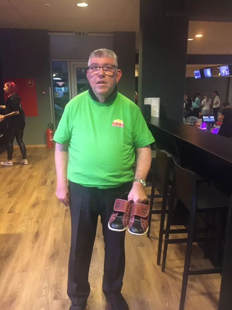 Homme en chemise verte tenant une paire de chaussures de bowling dans une piste de bowling.