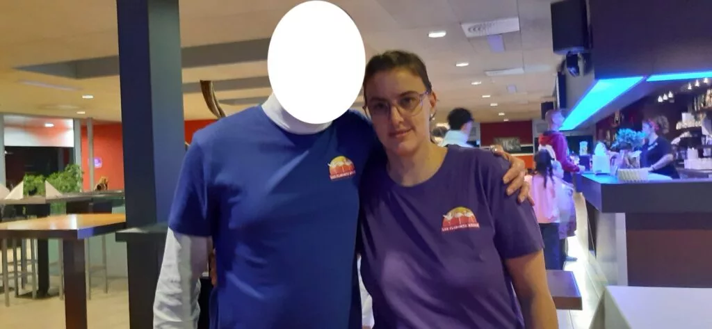 Deux personnes portant des t-shirts bleus et violets se tiennent côte à côte dans un bar. Le visage d'une personne est caché. Le bar est éclairé par un éclairage bleu.