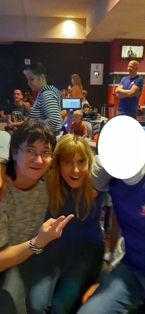 Deux personnes posant à l'intérieur avec un bras autour d'une personne dont le visage est caché. D'autres personnes et un arrière-plan coloré sont visibles.