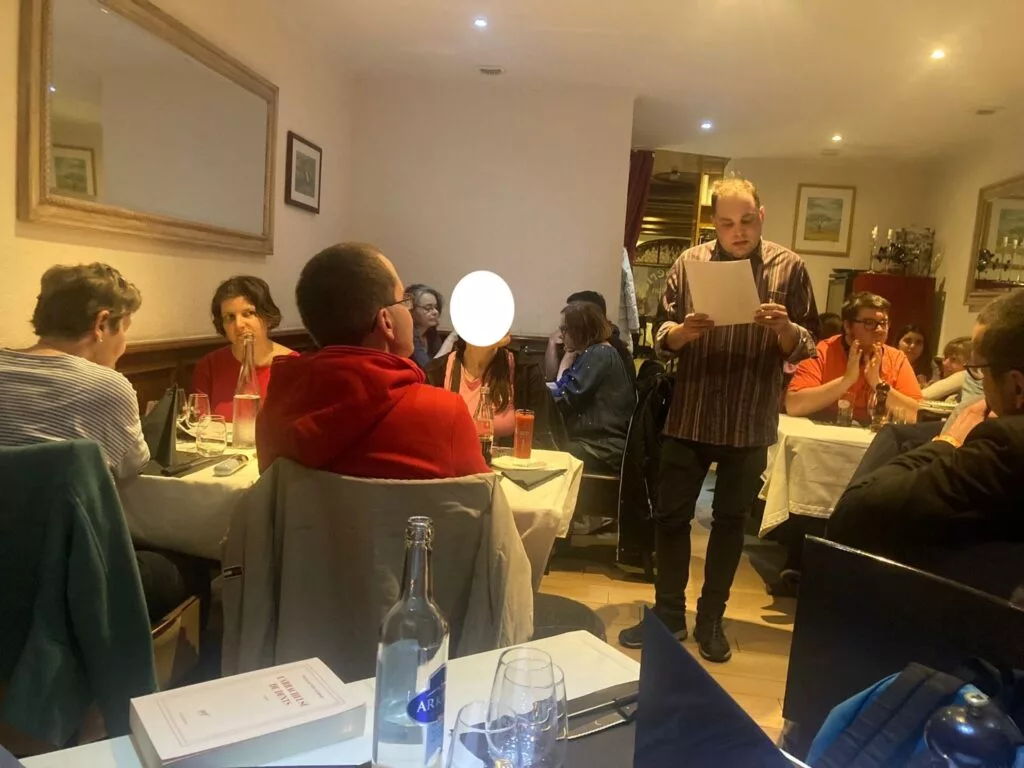 Une personne est debout et lit une feuille de papier dans un restaurant où des convives sont assis. Des verres et une bouteille d'eau sont disposés sur les tables. Les murs sont ornés d'œuvres d'art et d'un miroir.