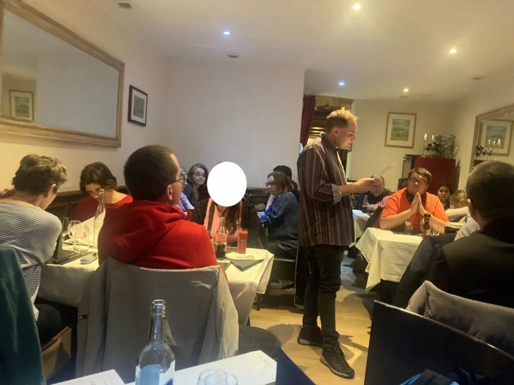 Scène de restaurant avec un serveur masculin prenant une commande auprès de clients assis à une table. Les murs sont décorés de photos encadrées. Les gens discutent et dînent.