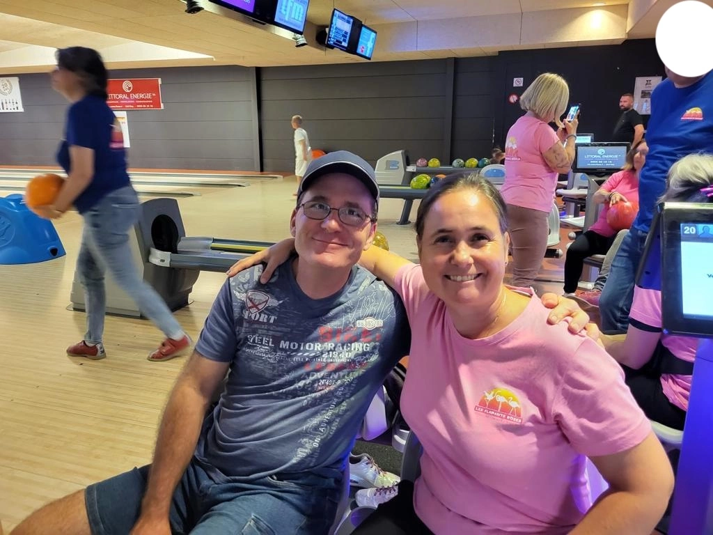 Deux individus assis dans une salle de bowling sourient à la caméra. L'un des deux passe son bras autour de l'autre. Des équipements de bowling et d'autres personnes sont visibles en arrière-plan.