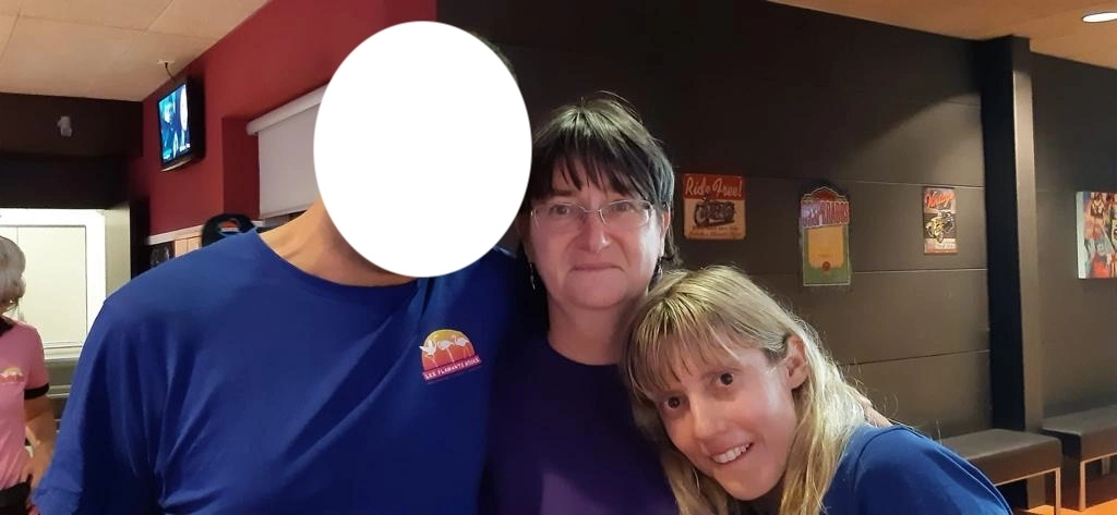 Trois personnes se tiennent debout à l'intérieur avec deux femmes au premier plan. Le visage de la personne à gauche est masqué. L'arrière-plan présente un mur avec des affiches.