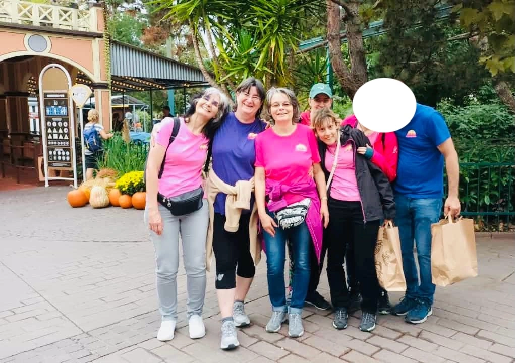 Groupe de six personnes posant à l'extérieur dans un parc. Cinq d'entre elles sont visibles, portant des vêtements décontractés. Deux d'entre elles tiennent des sacs de courses. L'arrière-plan présente des plantes et une entrée de bâtiment avec des citrouilles. Un visage est masqué.