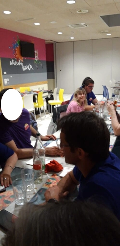 Un groupe de personnes assises autour d'une table dans un café aux couleurs vives. Plusieurs bouteilles et verres sont posés sur la table, avec une personne cachée.