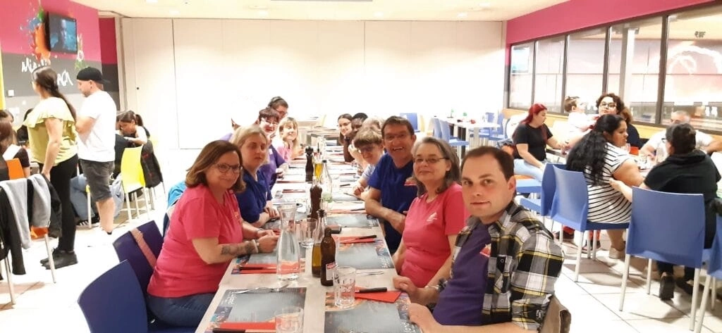 Un groupe de personnes assises à une longue table dans un restaurant, vêtues de vêtements décontractés. Certaines portent des chemises roses ou violettes. Des verres et des bouteilles sont posés sur la table. D'autres convives sont visibles en arrière-plan.