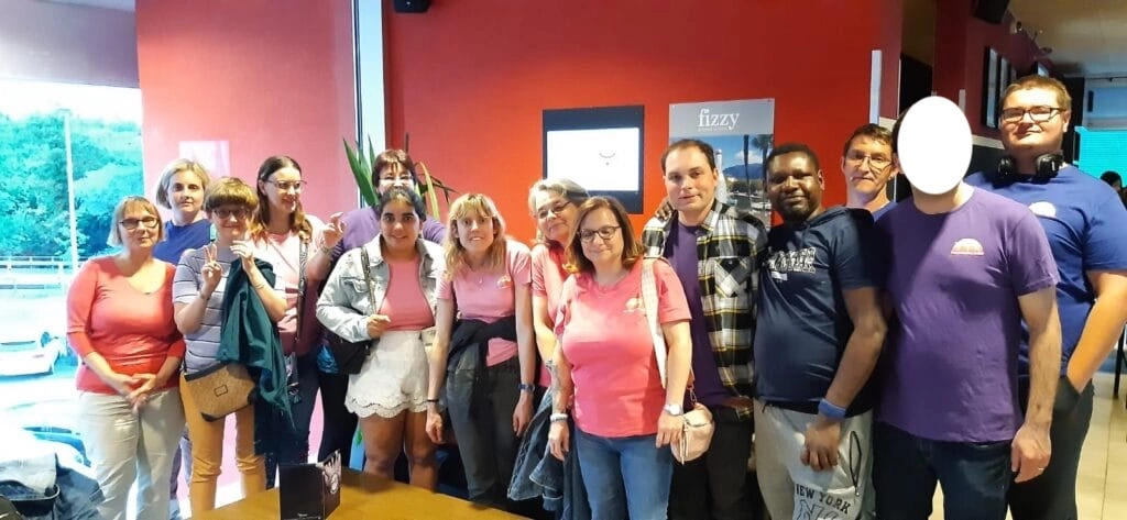 Un groupe diversifié de personnes pose à l'intérieur, vêtues de tenues décontractées, notamment de chemises roses et violettes. Un mur rouge et une fenêtre avec vue sur les arbres sont en arrière-plan.