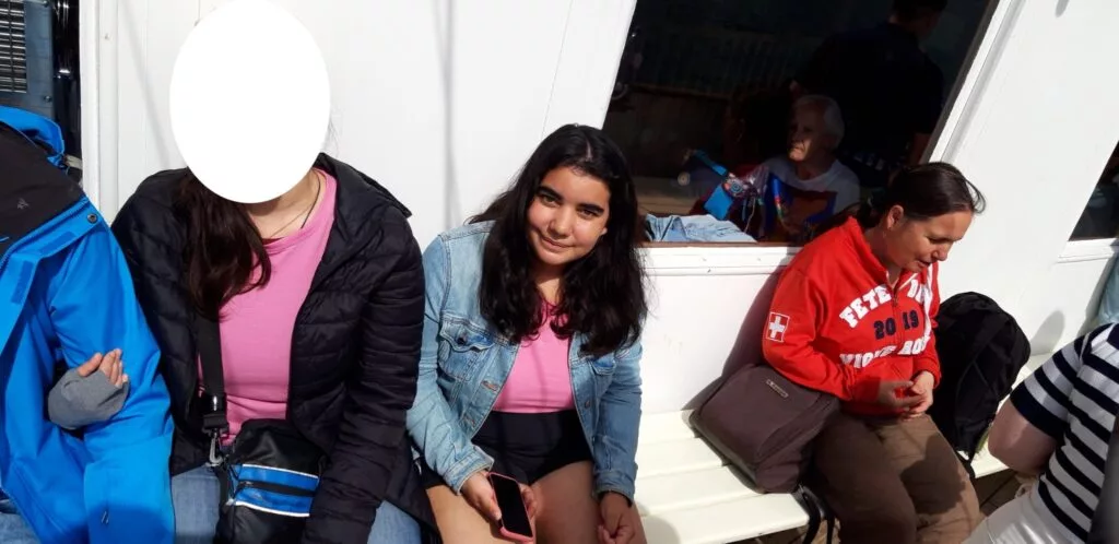 Trois personnes assises sur un banc ; l'une porte un haut rose et une veste noire, une autre porte une veste en jean et tient un téléphone, et une troisième porte un sweat-shirt rouge.