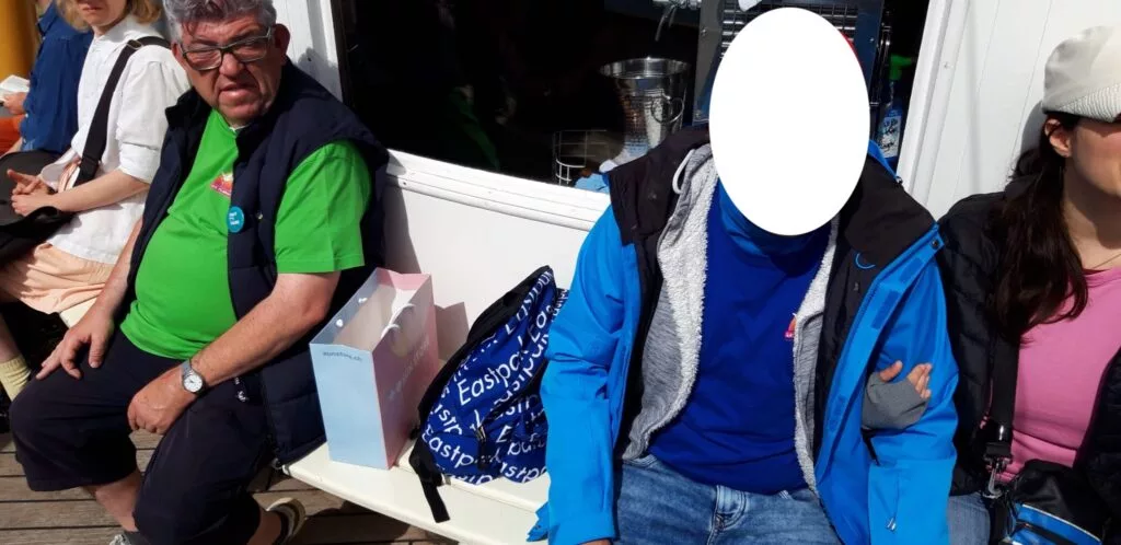 Trois personnes sont assises sur un banc à l'extérieur ; l'une d'elles a le visage caché. Des sacs de courses sont posés sur le banc.