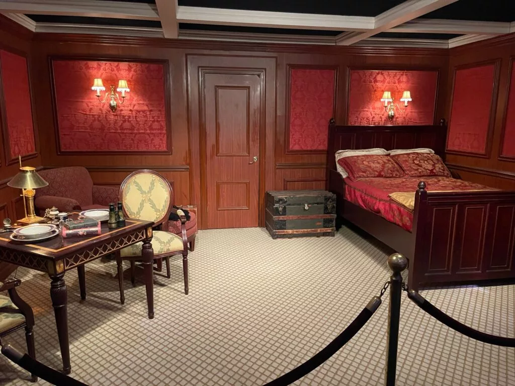 Cabine historique avec moquette rouge, lit en bois, table à manger et vieux coffre. La décoration comprend deux appliques murales et une lampe de table, toutes délimitées pour la vue.