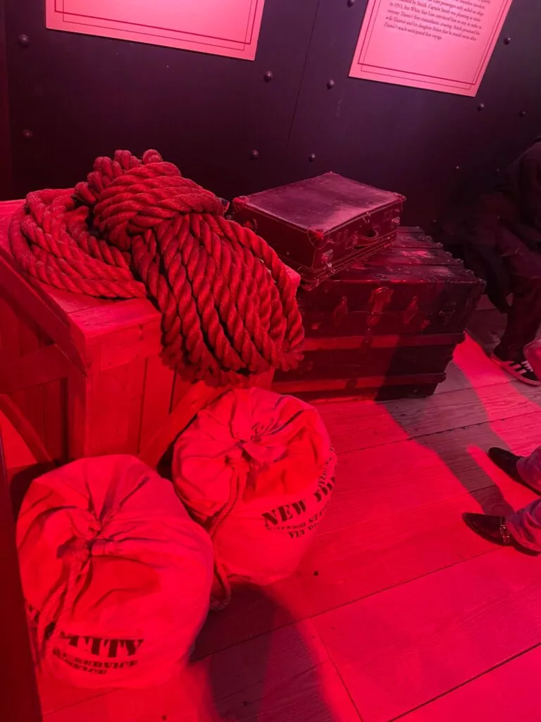 Scène éclairée en rouge avec des cordes enroulées sur une caisse en bois, deux sacs en toile de jute étiquetés « New Bedford » et une valise et une malle vintage sur un plancher en bois.