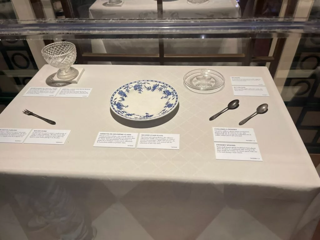 Vitrine présentant de la vaisselle historique, comprenant une assiette à motifs, deux cuillères, une fourchette, un plat en verre et un petit bol, chacun avec des étiquettes descriptives.