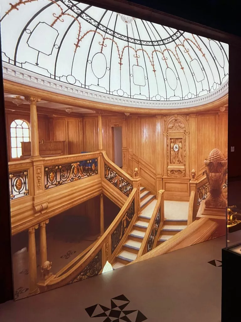 Escalier en bois complexe avec rampes ornées et une grande horloge sous un plafond en verre en forme de dôme.