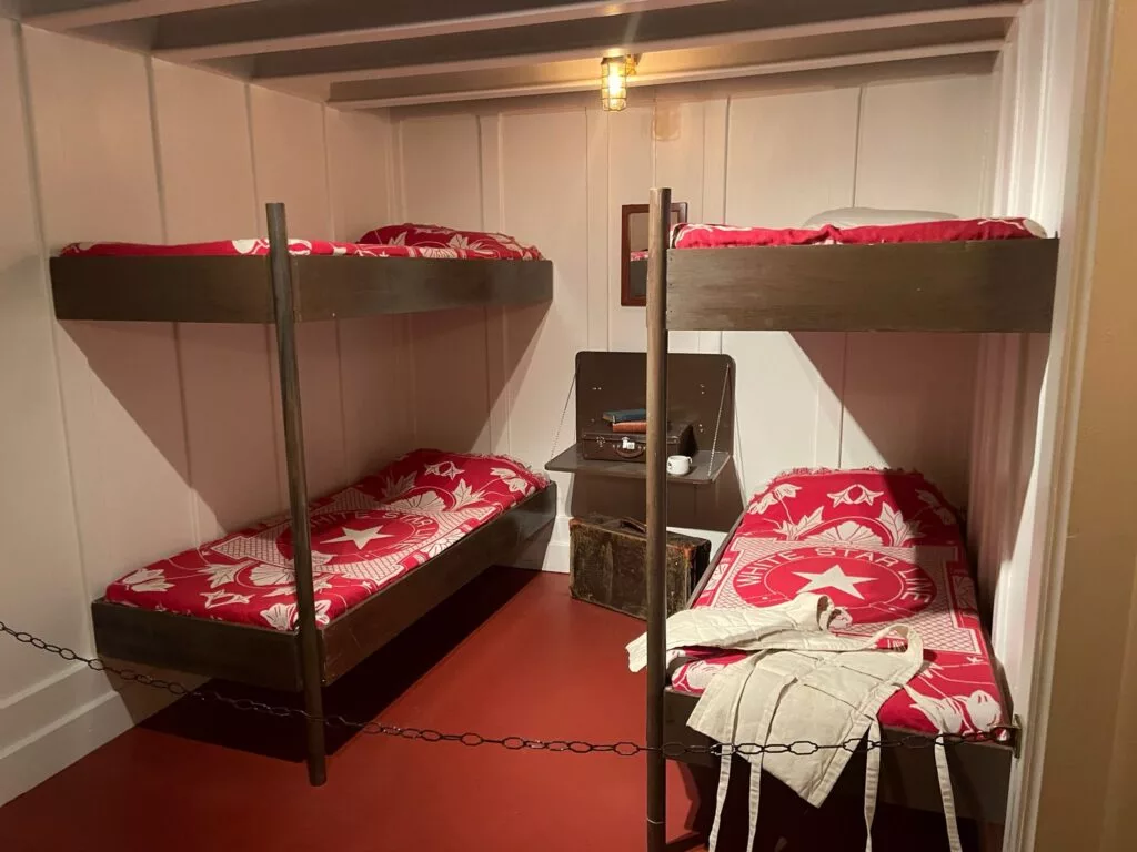 Une chambre avec deux lits superposés avec une literie à thème d'étoile rouge. Une petite table avec une valise, de la vaisselle et une lampe se trouve entre les lits. Une ampoule suspendue éclaire l'espace.