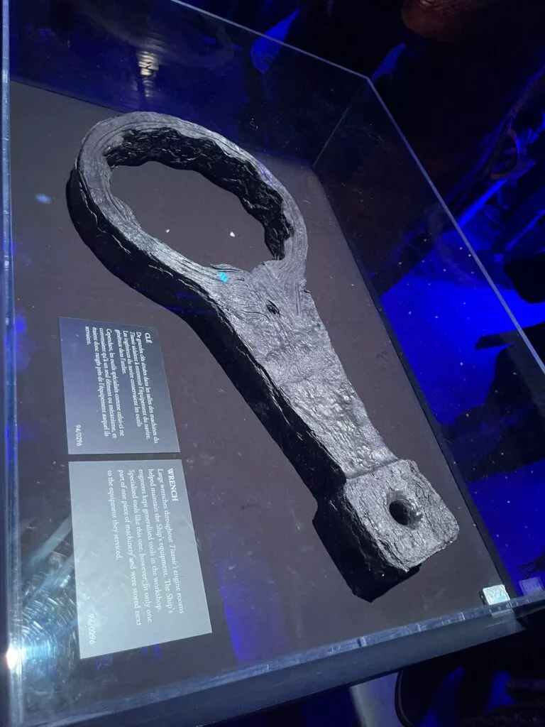 Une grande clé en fer rouillé exposée dans une vitrine en verre avec des plaques d'information, sous un éclairage bleu.