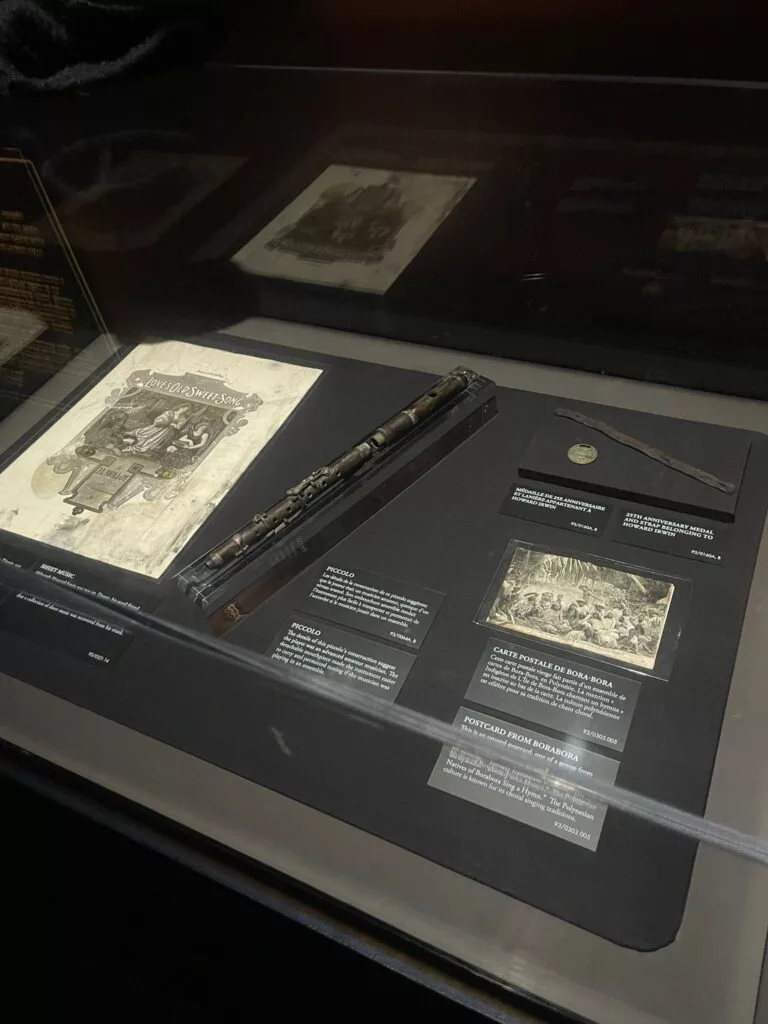 Vitrine contenant des documents historiques, un coffret en bois, une pièce de monnaie et un petit objet noir avec des étiquettes informatives sur fond sombre.