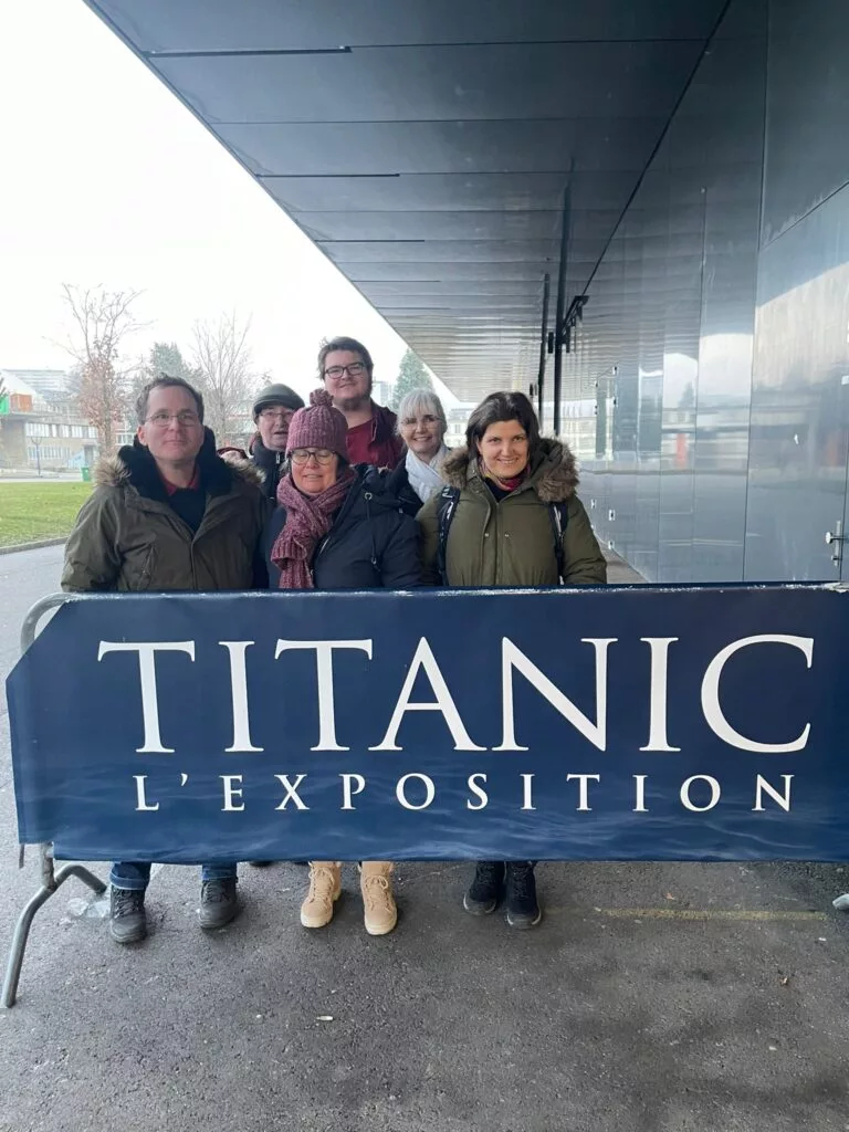 Un groupe de personnes debout derrière un panneau indiquant « Titanic L'Exposition » à l'extérieur d'un bâtiment moderne.