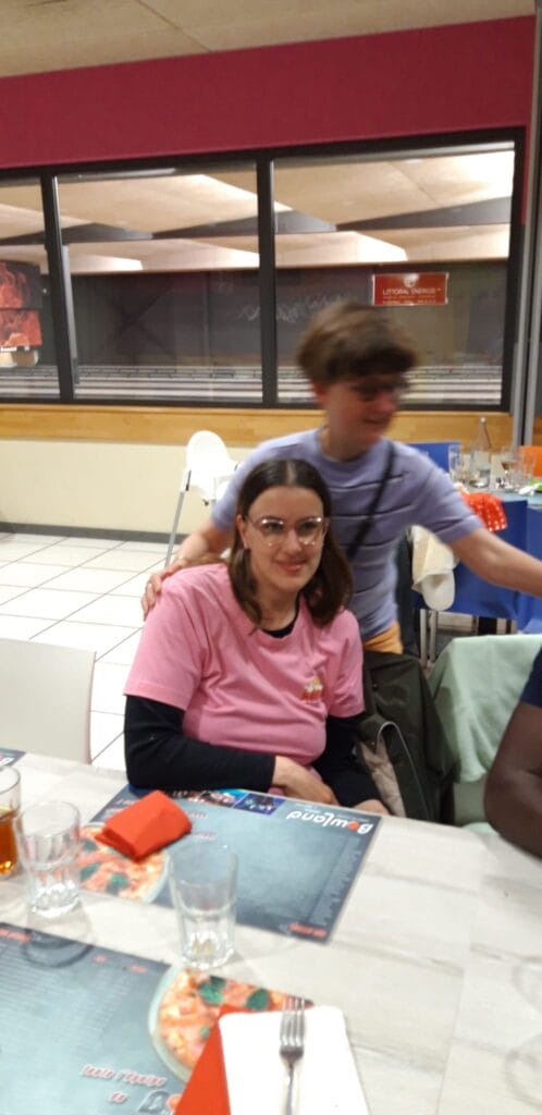 Une femme vêtue d'une chemise rose et de lunettes est assise à une table dans un restaurant. Une autre personne se tient derrière elle, légèrement floue. La table contient des menus, des boissons et d’autres articles de restauration.