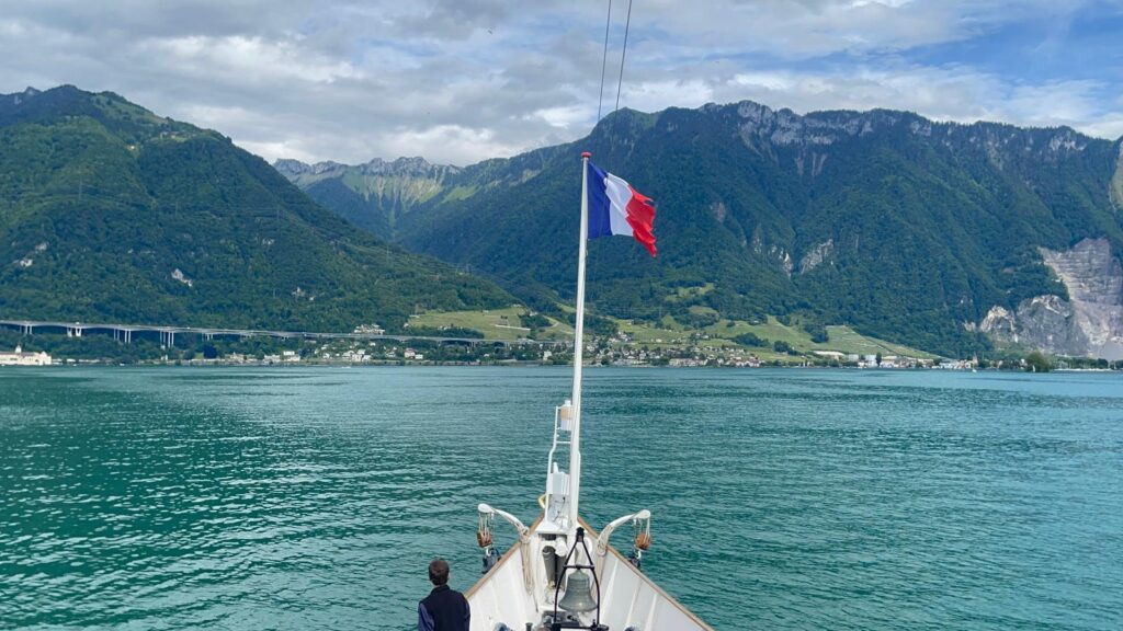 Bateau avec un drapeau français sur un lac, entouré de montagnes verdoyantes et d'un ciel partiellement nuageux.
