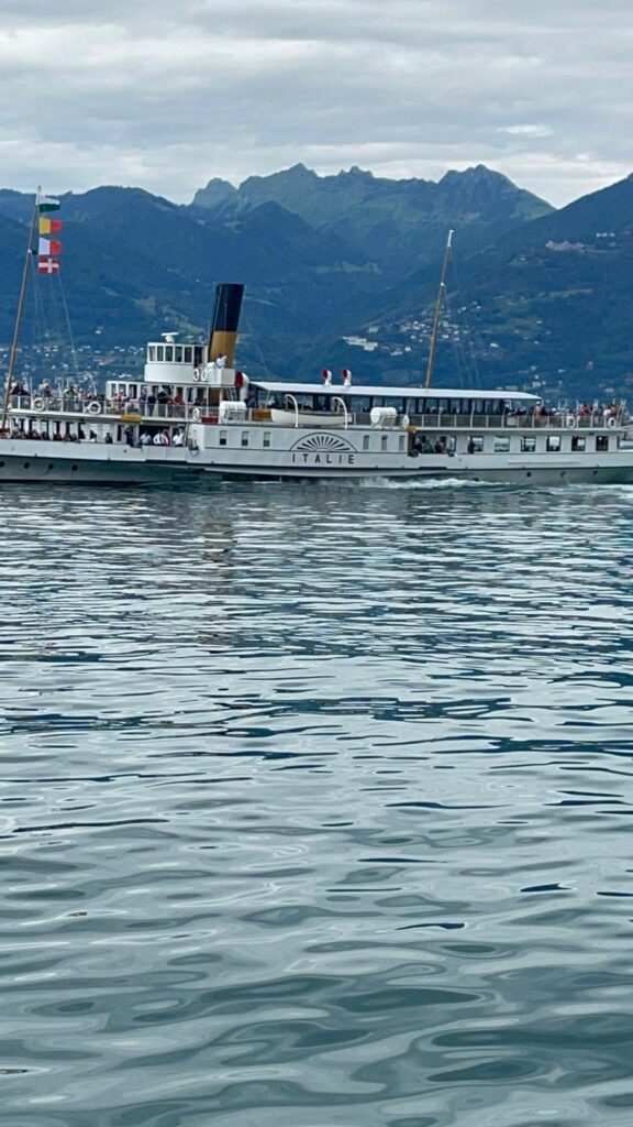 Un bateau à vapeur historique avec des drapeaux italiens navigue sur un lac calme, avec des montagnes en arrière-plan et un ciel nuageux au-dessus.
